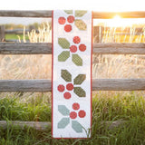 The boxed KT-17661 Holly-Day Cheer Runner Kit presented on a light ivory linen surface, showcasing the rich holiday red and spruce green fabric bundles inside.
